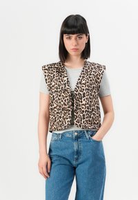 Vrouw met steil donker haar, gekleed in een mouwloos vest met panterprint over een lichtgrijs t-shirt en blauwe jeans, staand tegen een witte achtergrond.