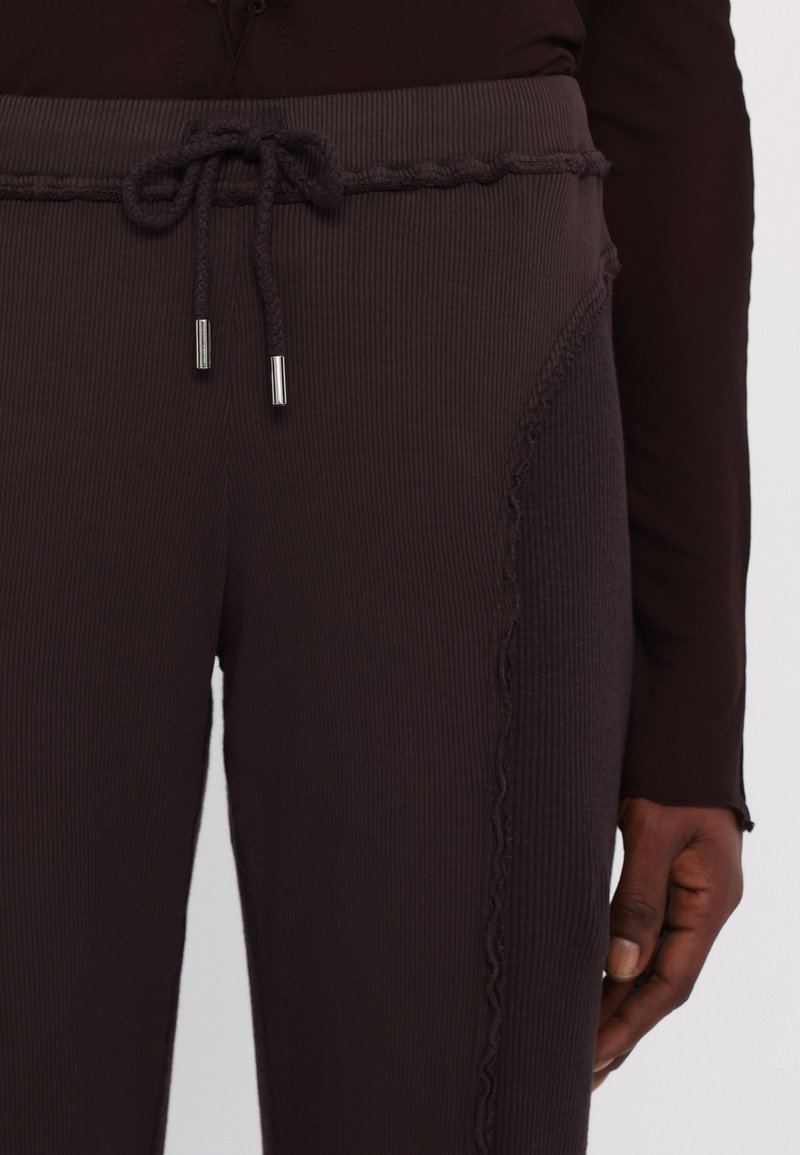 Pantalones deportivos oscuros de canalé con cintura elástica y cordón, puntas metálicas plateadas y costuras decorativas en los laterales. El material parece texturizado.