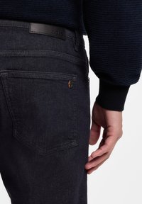 Jeans de mezclilla oscuro con una textura suave, que cuentan con una etiqueta de cuero negro con la marca en la cintura y costuras visibles en los bolsillos.