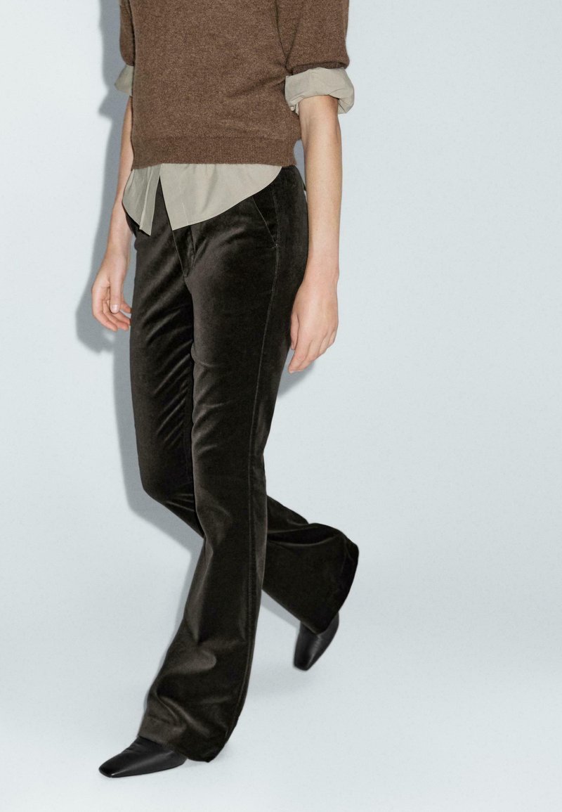 Brązowy wełniany sweter nałożony na jasną koszulę z kołnierzykiem, w połączeniu z ciemnymi welurowymi spodniami dzwonami i eleganckimi czarnymi butami.