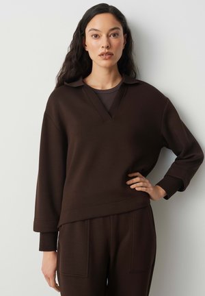 Vrouw met lang donker haar, gekleed in een donkerbruine trui en broek, staand met één hand op haar heup tegen een lichte achtergrond.