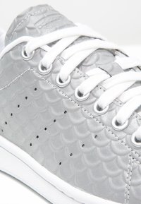 Zapatilla plateada con una textura escamada, cordones blancos y ojales redondos. Presenta una suela blanca sólida y pequeñas perforaciones negras.