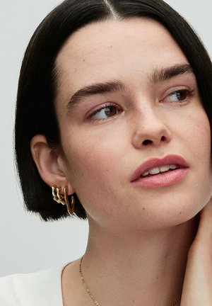 Goldfarbene Creolen zeigen ein organisches, verdrehtes Design. Glatte Oberfläche mit geschichteten Formen. Das Modell hat schulterlanges, dunkles Haar und dezentes Make-up.