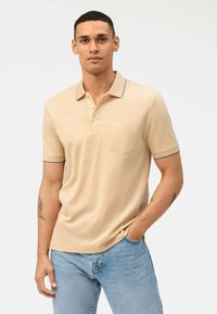 Beige poloshirt met een drieknopenlijn, detail van een zak en blauwe kraagaccenten, gecombineerd met lichtblauwe denim jeans.