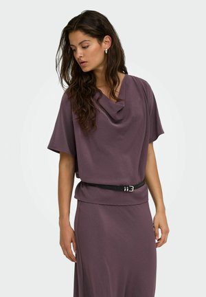Femme aux longs cheveux foncés portant un haut violet à manches courtes avec un col drapé et une jupe assortie avec une fine ceinture noire.