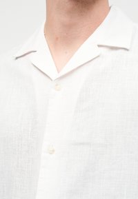 Άνδρας με λευκό λινό πουκάμισο με γιακά τύπου camp και δύο ορατά κουμπιά μπροστά.