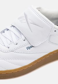 Λευκό δερμάτινο αθλητικό παπούτσι με αυτο�κόλλητο λουρί, υφασμάτινες πλάγιες πινελιές και καουτσούκ σόλα. Διαθέτει ασημένια σήμανση στο επάνω μέρος.