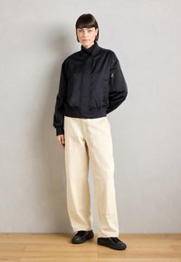 Veste bomber noire à col haut, poche zippée sur la manche, taille élastique, associée à un pantalon large beige clair et des chaussures noires.