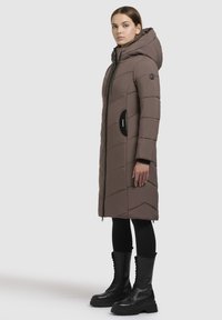 Langer, brauner Daunenmantel mit Kapuze, diagonaler Steppung, schwarzen Akzentreißverschlüssen und Seitentaschen, kombiniert mit schwarzen Schnürstiefeln.