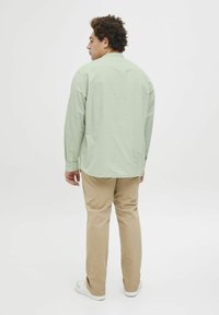 Camisa de manga larga verde claro con corte recto, puños abotonados y dobladillo curvado, combinada con pantalones beige y zapatillas blancas.