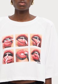 T-shirt blanc court avec un graphique de lèvres rouges brillantes avec une paille rayée, agencées en six carrés, avec le logo "FIORUCCO" en bas.