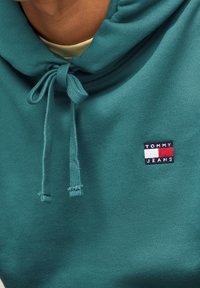 Felpa in cotone color teal con cappuccio e cordino, caratterizzata da una piccola toppa rettangolare con "TOMMY JEANS" nei colori navy, rosso e bianco. Texture liscia.
