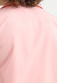 Světle růžová bunda vyrobená z hladké látky, na zadní straně nápis "NEW BALANCE" tučnými bílými písmeny zakřivený přes celou plochu. Červené akcenty na límci.