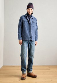 Berretto a righe blu scuro, giacca di jeans con colletto e due tasche sul petto, strato interno nero con zip, jeans blu chiaro e stivali marroni con lacci.