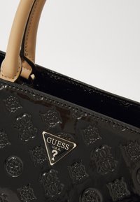 Juodas patentuotas odinis rankinukas, puošiamas įspaustais gėlių raštais, su béžinėmis rankenomis ir auksiniu trikampiu logotipu su "GUESS U.S.A."