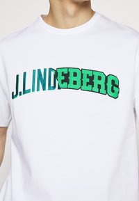 White cotton t-shirt featuring black and green embroidered text “J.LINDEBERG” in a bold, curved design across the chest.