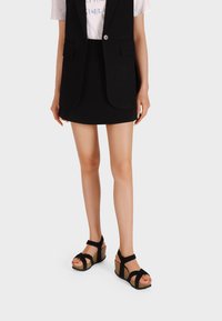 Blazer senza maniche nero, t-shirt bianca con grafica e mini gonna nera abbinati a sandali con zeppa neri con suole in sughero e cinturini incrociati.