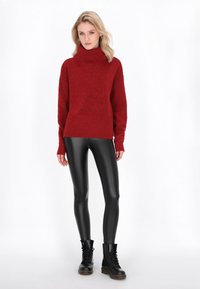 Roter gestrickter Rollkragenpullover mit geripptem Saum und Ärmeln, kombiniert mit glänzenden schwarzen Leggings und schwarzen Schnür-Stiefeletten.