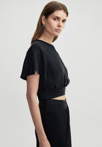 Zwarte cropped top met korte, fladderende mouwen, gemaakt van een gladde stof. Heeft een geplooid tailleband en een minimalistisch ontwerp.