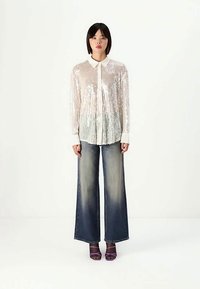 Una donna sta in piedi di fronte, indossando una camicia bianca trasparente con paillettes, jeans larghi e sbiaditi, e sandali con tacco viola su uno sfondo bianco semplice.