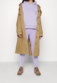 Gabardina beige con botones grandes sobre una sudadera ligera color púrpura y leggings a juego con rayas laterales. Zapatillas marrones con cordones.