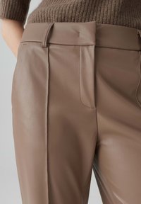 Bruine vegan leren broek met een gladde textuur, voorzien van een middelhoge tailleband, zijzakken en voorplooien.