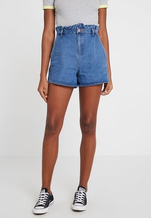 Vrouw draagt een blauwe spijkershort met hoge taille, een grijze geribbelde top met korte mouwen en gele rand, en zwarte lage sneakers met witte veters.