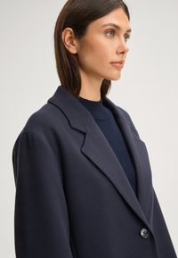 Navyblauwe oversized blazer met een notch-kraag, enkelknopen sluiting en zachte textuur. Gelaagd over een bijpassende geribbelde trui.
