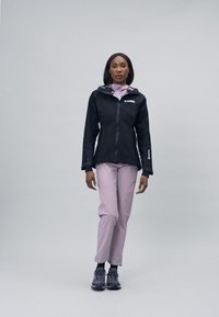Schwarze wasserdichte Jacke mit Kapuze, hellrosa Langarmshirt, rosa Hose und graue Sportschuhe, mit elegantem Design und funktionalen Elementen.
