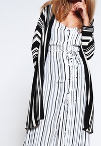 Svart och vit randig lång cardigan med öppen front och vida ärmar över en vitrandig byxdress med knappar och knytning i midjan.