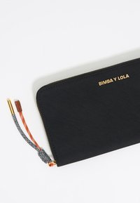 Mustast riidest rahakott, millel on kuldne "BIMBA Y LOLA" logo, tekstuuriga aktsendid ja värvilised kangast tõmblukud.