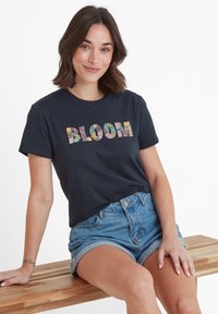 Woman with shoulder-length brown hair sits on wooden bench wearing navy T-shirt with "BLOOM" floral text and rolled denim shorts.