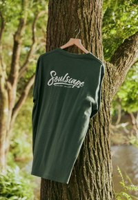 T-shirt à manches longues vert olive suspendu à un cintre en bois, avec un logo « Soulscape » en cursive blanche au dos. Tissu en coton, coupe décontractée.