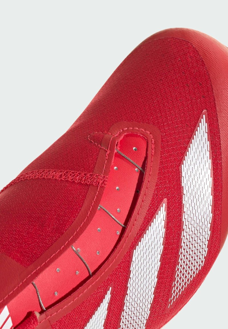 Roter Sportschuh mit einer strukturierten Oberseite, ausgestattet mit weißen Akzenten und Netzpaneelen. Das Design umfasst Schnürsenkel und metallische Details für Stabilität.