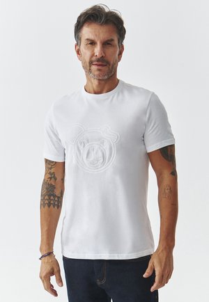 Tricou alb din bumbac cu un design texturat al feței unei urși pe partea din față. Mâneci scurte și gât rotund. Material neted, stil ajustat.