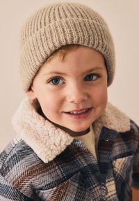 Bonnet en tricot côtelé beige sur un enfant. L'enfant porte une veste à carreaux bleus et bruns avec un col en fausse fourrure crème. Peau lisse et cheveux clairs.