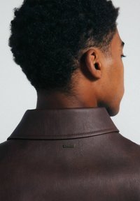 Braune Lederjacke mit einem Reverskragen, sichtbaren Nähten und einem metallischen Logosticker auf der Rückseite in Nackenhöhe.