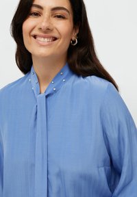 Blusa azzurro chiaro con colletto a fiocco, caratterizzata da un tessuto testurizzato, una leggera lucentezza e perle decorative lungo il colletto.