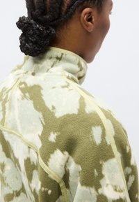 Ζακέτα φλις σε πράσινο και κρεμ μοτίβο τι-νταϊ, με ψηλό γιακά, ραφές αντίθεσης και απαλή υφή.