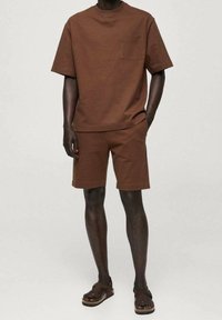 Homme portant une chemise brune à manches courtes avec une poche poitrine, un short assorti et des sandales ouvertes marron, se tenant devant un fond uni.