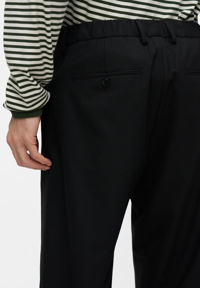 Personne portant un pantalon noir avec une poche arrière boutonnée et une chemise à manches longues rayée verte et blanche.