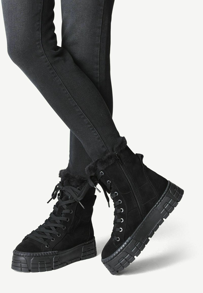 Bottines noires à lacets avec semelles épaisses en caoutchouc et bordure en fourrure, portées avec un pantalon noir ajusté.