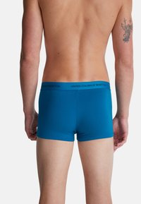Torso e gambe maschili di spalle, che indossano boxer blu con elastico "United Colors of Benetton" e un tatuaggio di angelo sul braccio destro.