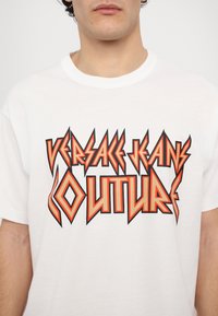 Tricou alb din bumbac, cu un text grafic îndrăzneț în roșu și negru "VERSACE JEANS COUTURE" într-un font unghiular. Design cu mâneci scurte și guler rotund.