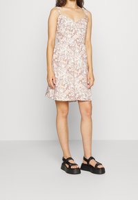 Žena v bezrámovém šatech po kolena s růžovým a bílým paisley vzorem a černými platformovými sandály, stojící před jednobarevným pozadím.