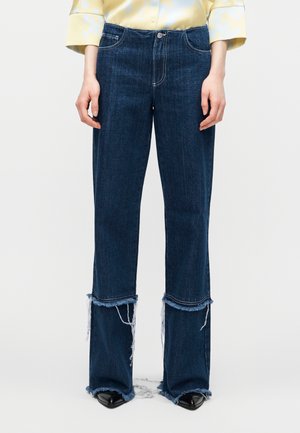 Donkere blauw wijd uitlopende jeans met rafelige zoom en uitgesneden details boven de enkels, gedragen met zwarte puntschoenen en een lichtgeel shirt.