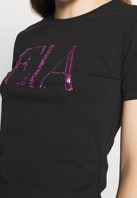 Svart t-shirt i bomull med en stor, paljetterad rosa logotyp "EA" och texten "Emporio Armani". Åtsittande med rundad halsringning.