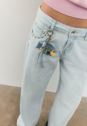 Persona che indossa jeans azzurri con borchie e un portachiavi a forma di pesce attaccato a una passante della cintura, e un top corto rosa in maglia.