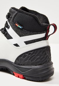 Nilkkakorkeinen saapas, jossa on teksturoitu musta päällinen, valkoiset yksityiskohdat sekä kova kumipohja, jossa on punaisia ja mustia yksityiskohtia pitoa ja tukea varten.