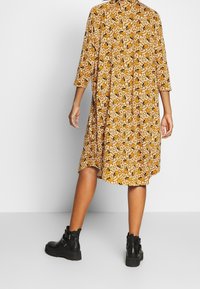 Dlouhé žluté šaty s květinovým vzorem, tři čtvrtinové rukávy, volný střih a lem, který sahá pod kolena; nošeny s černými kotníkovými botami.